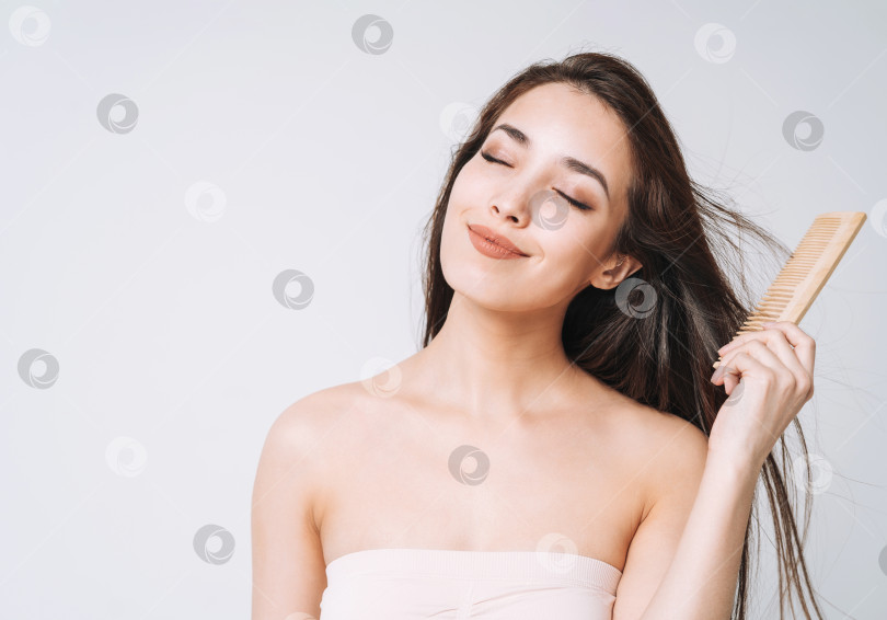 Скачать Красивый портрет счастливой улыбающейся азиатской женщины с темными длинными волосами, расчесывающей деревянным гребнем на белом фоне изолированно фотосток Ozero