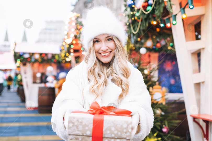 Скачать Молодая счастливая женщина блондинка с вьющимися волосами в белой меховой шапке с подарочной коробкой в руках на рождественской ярмарке на зимней улице, украшенной огнями фотосток Ozero