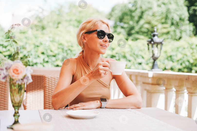 Скачать Взрослая элегантная блондинка в солнцезащитных очках и бежевой одежде с чашкой кофе в уличном кафе летнего города фотосток Ozero