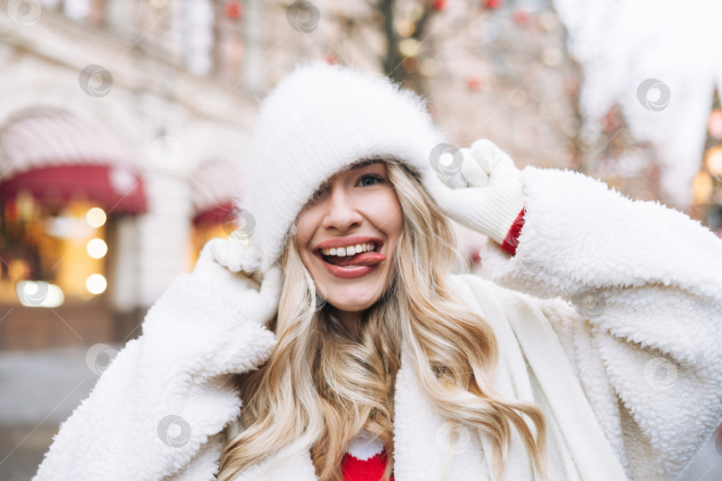 Скачать Молодая счастливая женщина с вьющимися волосами в белой меховой шапке веселится на рождественской ярмарке на зимней улице, украшенной огнями фотосток Ozero