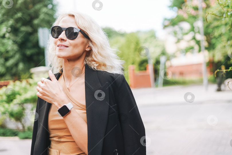 Скачать Взрослая элегантная блондинка в черном жакете и солнцезащитных очках на улице летнего города фотосток Ozero