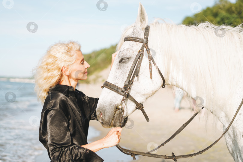 Скачать Молодая блондинка в черной одежде верхом на белой лошади на фоне морского пейзажа фотосток Ozero