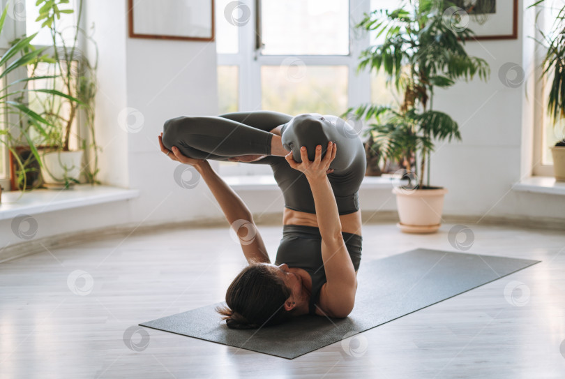 Скачать Молодая подтянутая женщина практикует йогу, выполняя асаны в легкой студии йоги с зеленым комнатным растением фотосток Ozero