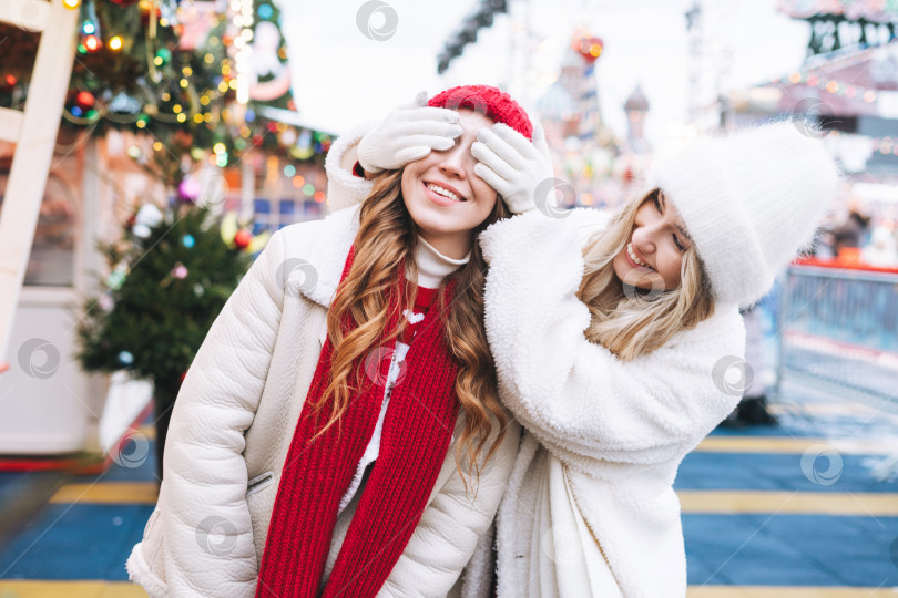 Скачать Молодые счастливые подруги с вьющимися рыжими волосами веселятся на зимней улице, украшенной огнями фотосток Ozero