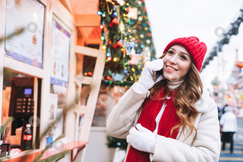 Скачать Молодая счастливая женщина с вьющимися волосами в красной вязаной шапочке пользуется мобильным телефоном на рождественской ярмарке на зимней улице, украшенной огнями фотосток Ozero