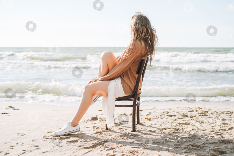 Скачать Молодая беззаботная красивая женщина с длинными волосами в свитере сидит на стуле на морском пляже фотосток Ozero