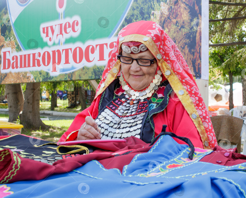 Скачать Пожилая женщина в башкирской одежде вышивает крючком. Традиционный национальный праздник Сабантуй в городском парке. фотосток Ozero