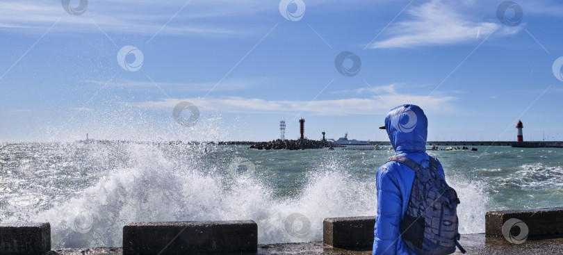 Скачать Мужчина с рюкзаком на морской набережной, смотрит на сильные волны, брызги воды и маяк. фотосток Ozero