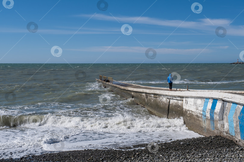 Скачать Зимний морской пейзаж. Девушка в синей куртке стоит на волнорезе и смотрит на волны. фотосток Ozero