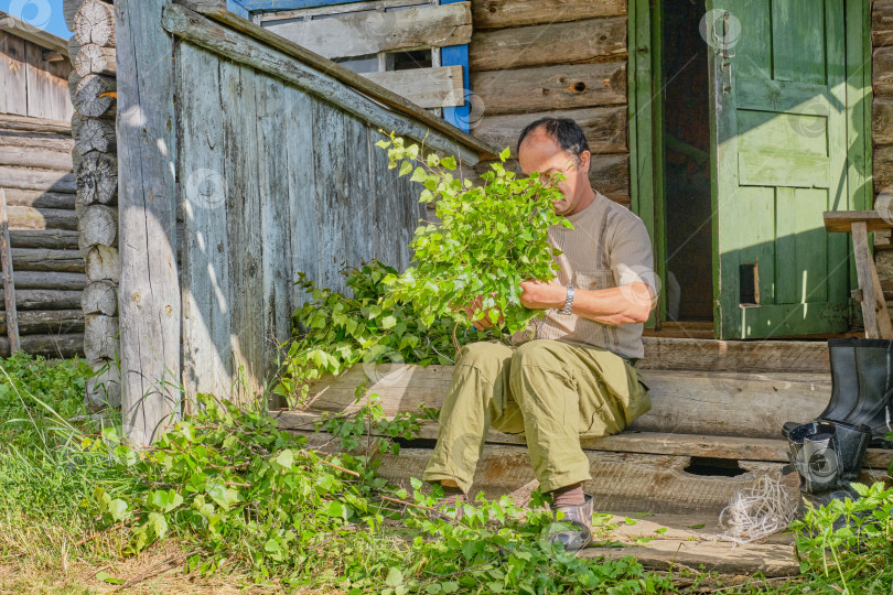 Скачать Мужчина связывает березовые веники для бани, сидя на деревянном крыльце старого загородного дома в сибирской деревне, Россия фотосток Ozero