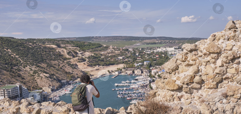 Скачать Вид на Балаклавскую бухту, город Севастополь, Крымский полуостров, Россия. Турист-мужчина с рюкзаком фотографирует на фотоаппарат фотосток Ozero