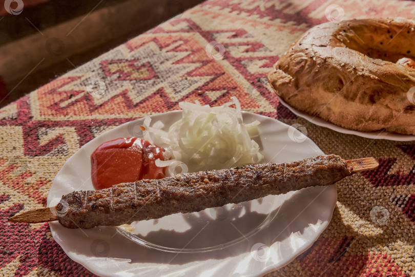 Скачать Люля-кебаб из баранины с томатным соусом и маринованным луком на белой тарелке. На столе скатерть с красным азиатским узором. фотосток Ozero