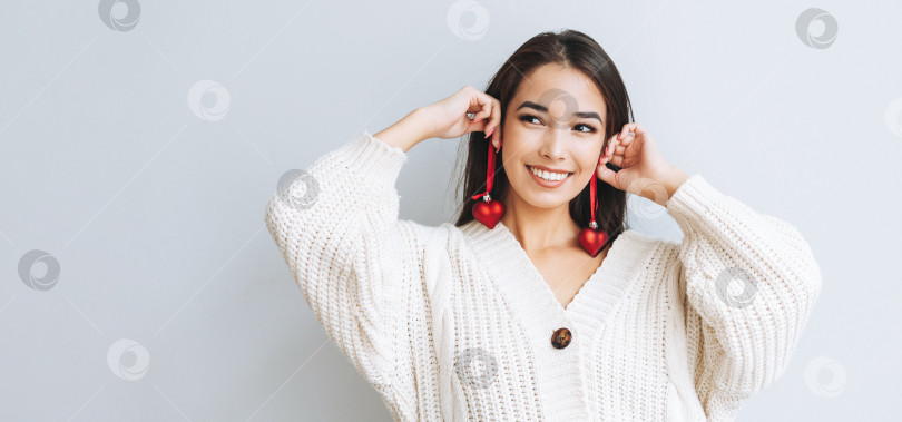 Скачать Молодая счастливая азиатская девушка с длинными волосами в белом вязаном кардигане с рождественским шаром в виде красного сердечка в руках на сером фоне, зимнее рождество фотосток Ozero