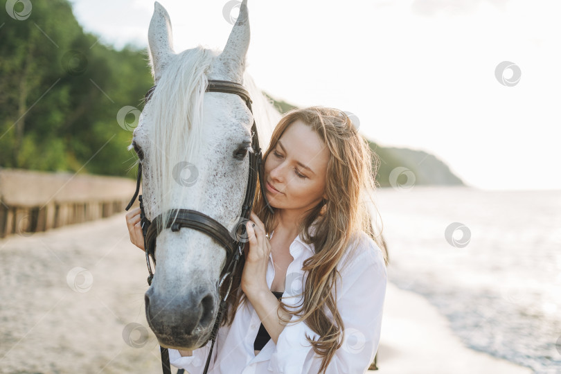 Скачать Молодая длинноволосая женщина в белой рубашке с белой лошадью на фоне морского пейзажа фотосток Ozero