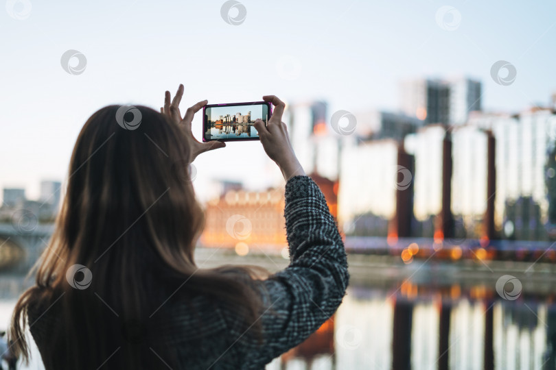 Скачать Молодая женщина в пальто фотографирует на смартфон вечерний городской пейзаж фотосток Ozero