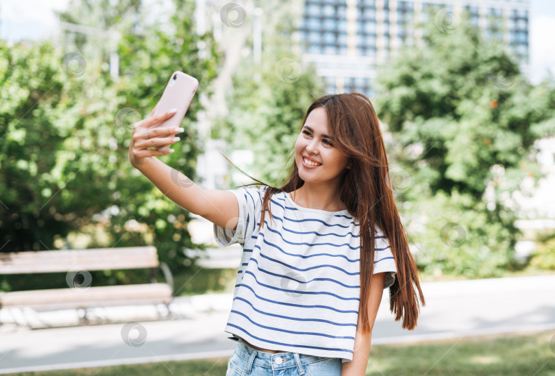 Скачать Портрет молодой студентки с длинными волосами, делающей селфи с помощью мобильного телефона в городском парке фотосток Ozero