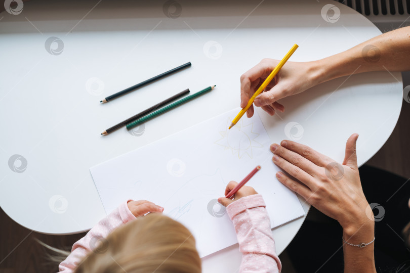 Скачать Маленькая девочка-малыш со своей мамой рисуют цветными карандашами на столе в детской комнате дома, вид сверху фотосток Ozero