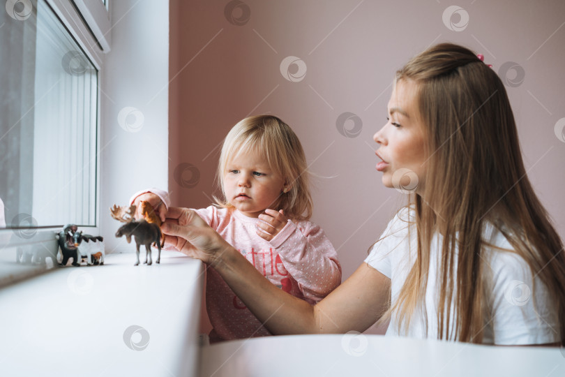 Скачать Маленькая девочка-малыш со своей матерью играют с игрушками в виде животных на подоконнике в детской комнате дома фотосток Ozero