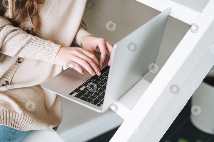 Скачать Обрезанная фотография молодой женщины с длинными волосами в кардигане, работающей за ноутбуком, сидя на лестнице дома фотосток Ozero