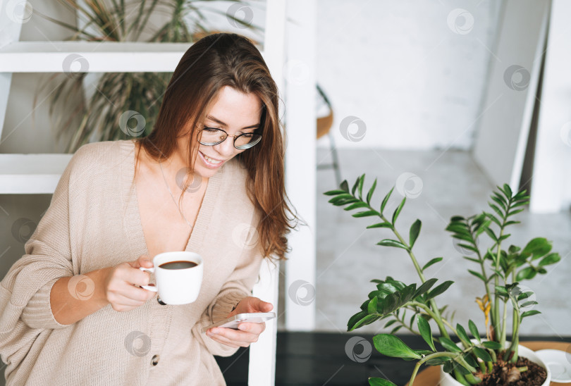 Скачать Улыбающаяся женщина с длинными волосами в бежевом кардигане пьет кофе и пользуется мобильным телефоном дома фотосток Ozero