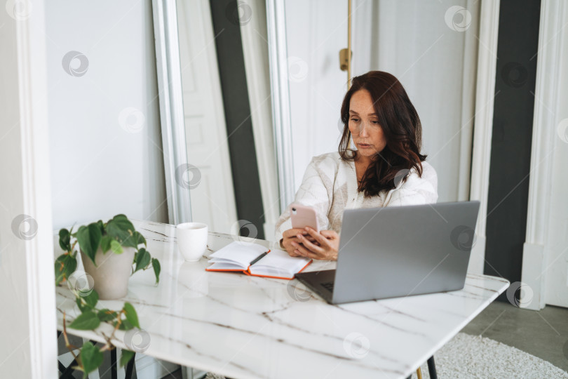 Скачать Думающая брюнетка с длинными волосами в белом кардигане работает на ноутбуке с помощью мобильного телефона в светлом современном офисе фотосток Ozero