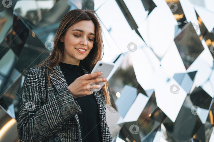 Скачать Молодая улыбающаяся женщина в пальто с чашкой кофе, пользующаяся мобильным телефоном на улице вечернего города фотосток Ozero
