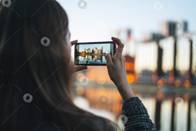 Скачать Молодая женщина в пальто фотографирует на смартфон вечерний городской пейзаж фотосток Ozero