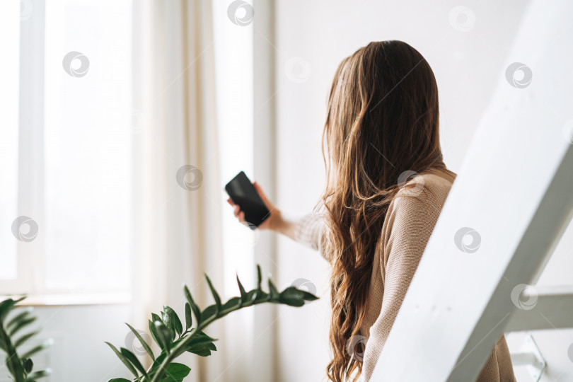 Скачать Молодая женщина с длинными волосами брюнетки в уютном вязаном кардигане пользуется мобильным телефоном в ярком интерьере дома фотосток Ozero