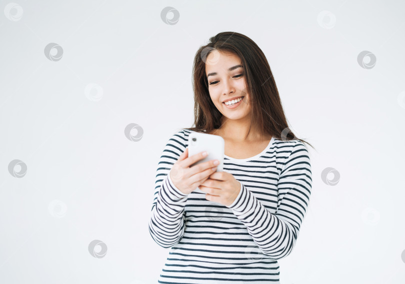 Скачать Молодая счастливая азиатка с длинными волосами в полосатом лонгсливе, использующая мобильный телефон на белом фоне изолированно фотосток Ozero