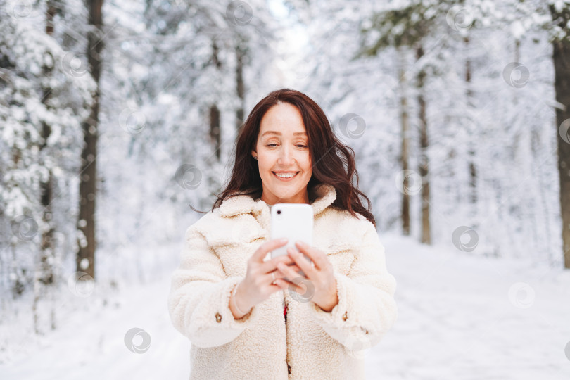 Скачать Улыбающаяся женщина с темными волосами в зимней одежде, пользующаяся мобильным телефоном на фоне заснеженного зимнего леса фотосток Ozero