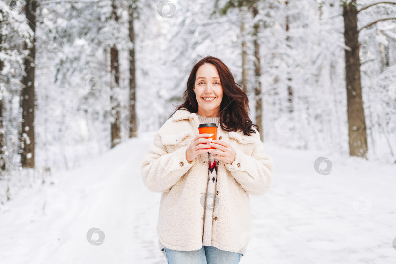 Скачать Улыбающаяся женщина с темными волосами в зимней одежде с бумажным стаканчиком кофе в руках на фоне заснеженного зимнего леса фотосток Ozero