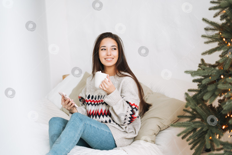 Скачать Молодая красивая азиатка с темными длинными волосами в уютном свитере пользуется мобильным смартфоном на кровати в комнате с рождественской елкой дома фотосток Ozero