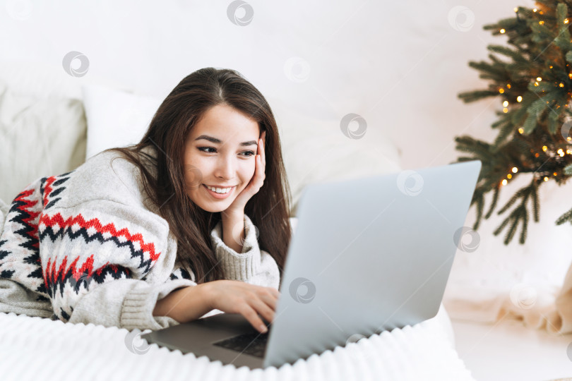 Скачать Молодая азиатка в уютном свитере пользуется ноутбуком на кровати в комнате с рождественской елкой дома фотосток Ozero
