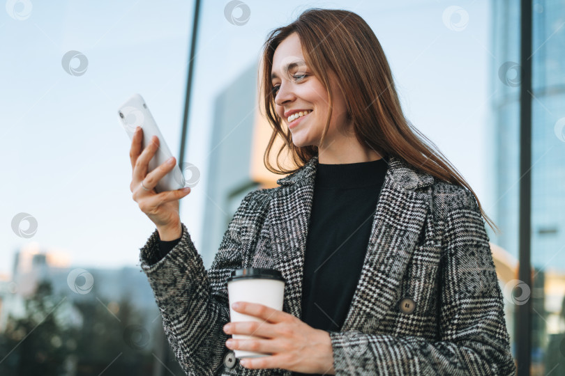 Скачать Молодая улыбающаяся женщина в пальто с чашкой кофе, пользующаяся мобильным телефоном на улице вечернего города фотосток Ozero