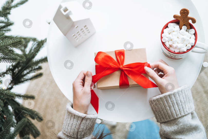 Скачать Молодая женщина в уютном свитере с подарочной коробкой с красной лентой в комнате с рождественской елкой дома фотосток Ozero