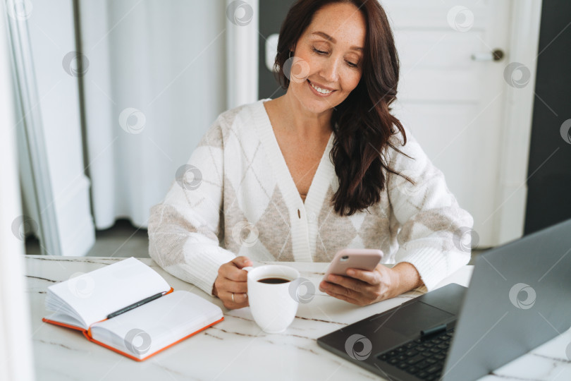 Скачать Улыбающаяся брюнетка с длинными волосами в белом кардигане работает на ноутбуке с помощью мобильного телефона в светлом современном офисе фотосток Ozero