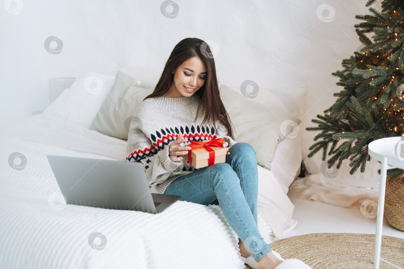Скачать Молодая азиатка в уютном свитере пользуется ноутбуком на кровати в комнате с рождественской елкой дома фотосток Ozero