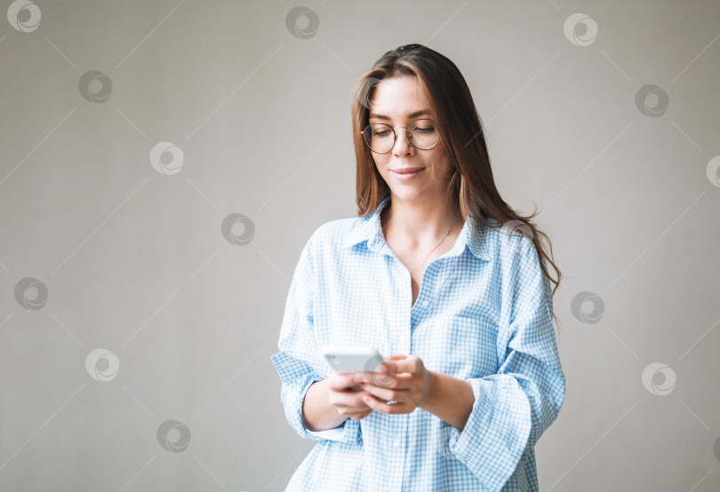 Скачать Портрет молодой улыбающейся женщины с темными длинными волосами в синей рубашке, использующей мобильный телефон в руке на сером фоне фотосток Ozero