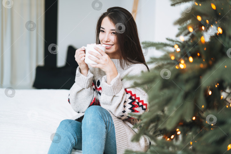 Скачать Молодая женщина в уютном свитере с чашкой какао с зефиром в комнате с рождественской елкой дома фотосток Ozero