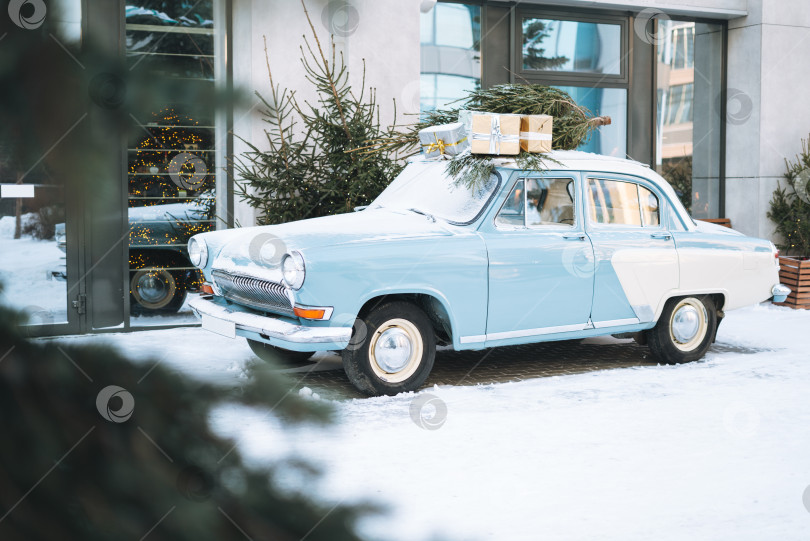 Скачать Винтажный светло-голубой автомобиль, украшенный натуральной рождественской елкой и коробками с подарками на зимней улице фотосток Ozero