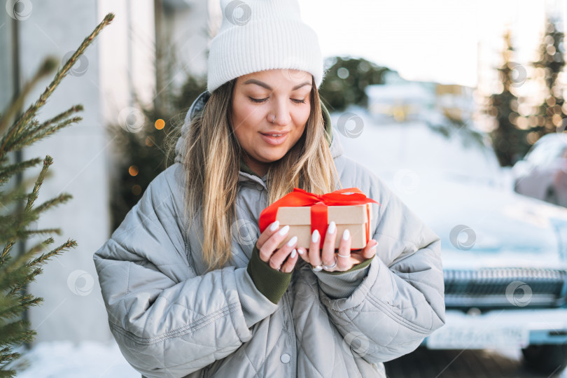 Скачать Молодая счастливая женщина-блондинка в теплой одежде с подарочной коробкой в руках возле ретро-автомобиля, украшенного рождественской елкой на зимней улице фотосток Ozero