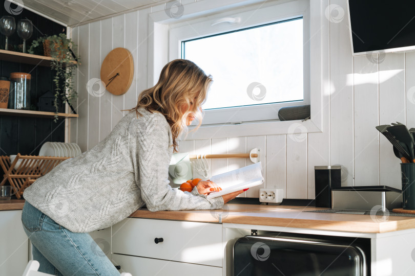 Скачать Молодая женщина со светлыми вьющимися волосами в сером свитере читает книгу на кухне дома фотосток Ozero