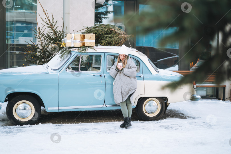 Скачать Молодая женщина-блондинка в теплой одежде с чашкой кофе в руках возле ретро-автомобиля, украшенного рождественской елкой на зимней улице фотосток Ozero
