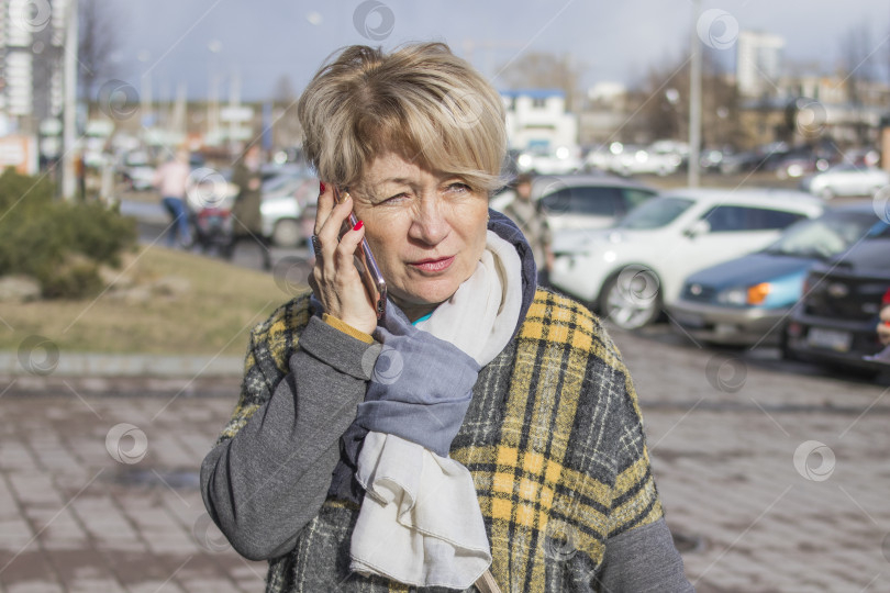 Скачать Симпатичная пожилая женщина в повседневной одежде разговаривает по телефону на весенней городской улице. фотосток Ozero