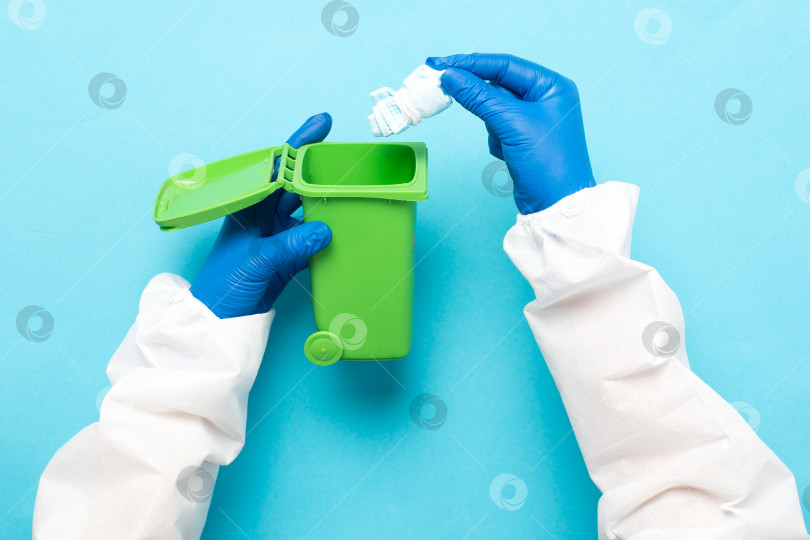 Скачать Выбрасываем медицинскую маску в зеленый контейнер для вторичной переработки на синем фоне. Синие маски для лица после использования. Использование медицинских масок фотосток Ozero