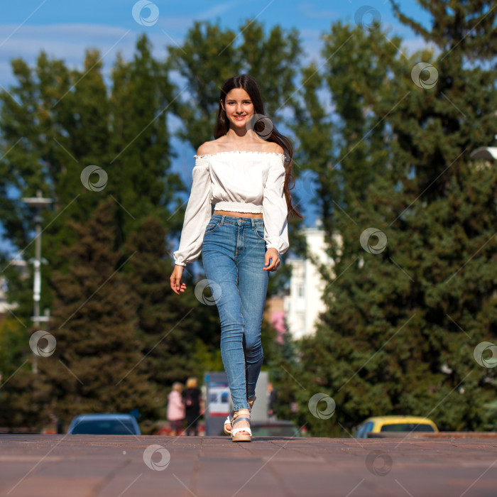 Скачать Молодая красивая брюнетка в джинсах и белой блузке прогуливается по летней улице фотосток Ozero