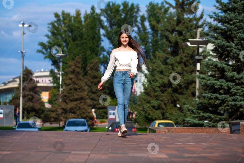 Скачать Молодая красивая брюнетка в джинсах и белой блузке прогуливается по летней улице фотосток Ozero