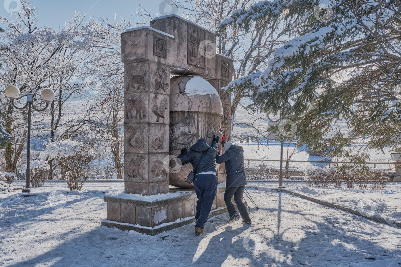 Скачать Мужчина и женщина в повседневной теплой одежде переворачивают яйцо желаний. Курортный парк зимой. Каменные скульптурные солнечные часы со знаками зодиака фотосток Ozero