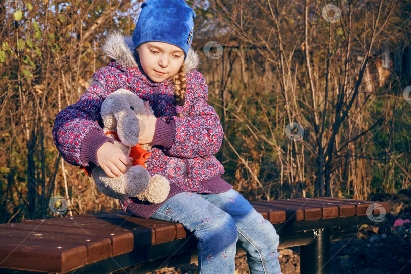 Скачать Девочка сидит на скамейке в парке и обнимает плюшевого кролика. Место для прогулок и отдыха фотосток Ozero