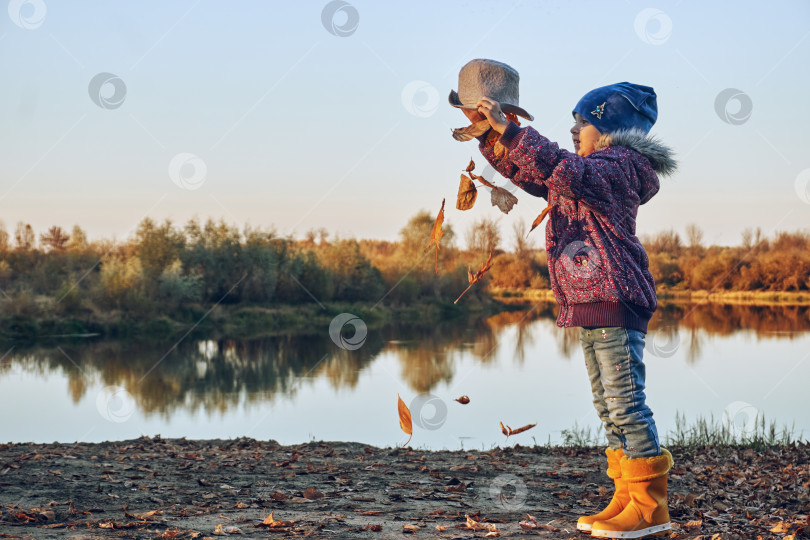 Скачать Девочка, высыпающая сухие листья из своей шляпы, игры и развлечения на свежем воздухе, осень. Песчаный берег фотосток Ozero